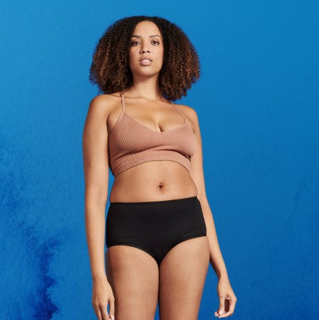 Woman standing, wearing a brown tank top and black Soft Cotton washable incontinence underwear, against a blue background.