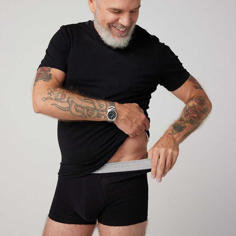 TENA Men Protective Boxers Light: Man in black shirt and grey underwear with tattoos, showcasing washable, discreet incontinence protection, designed for comfort and everyday use.