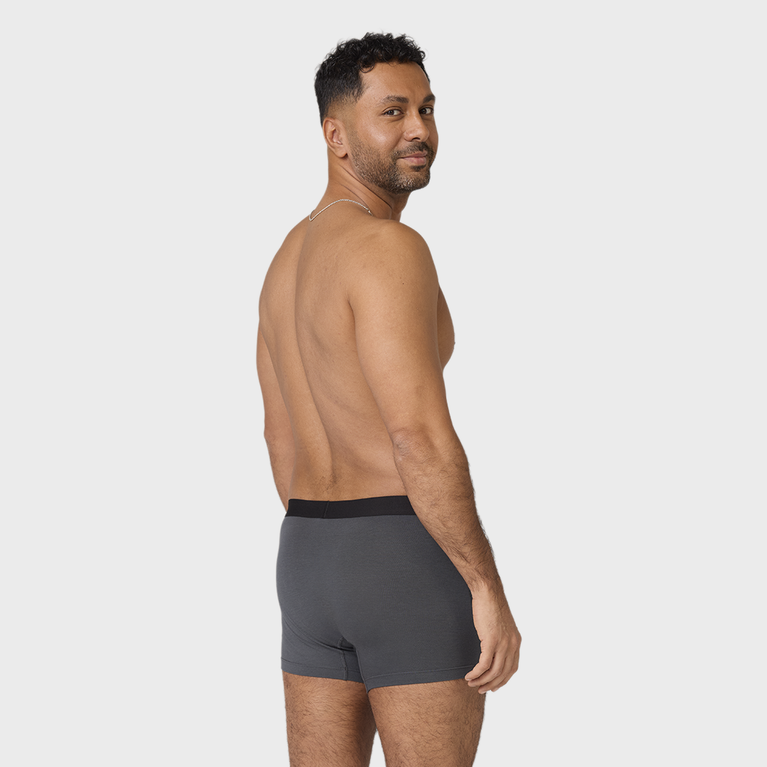 Man in dark gray NEW TENA Men Protective Boxers, glancing back, showcasing sleek design and fit with a silver chain against a light gray background.