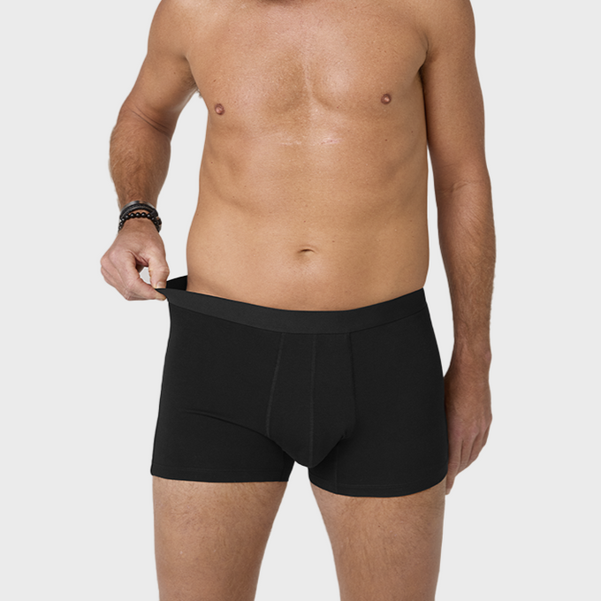 Man stands confidently wearing black TENA Men Washable Incontinence Protective Boxers, posing against a plain grey background.