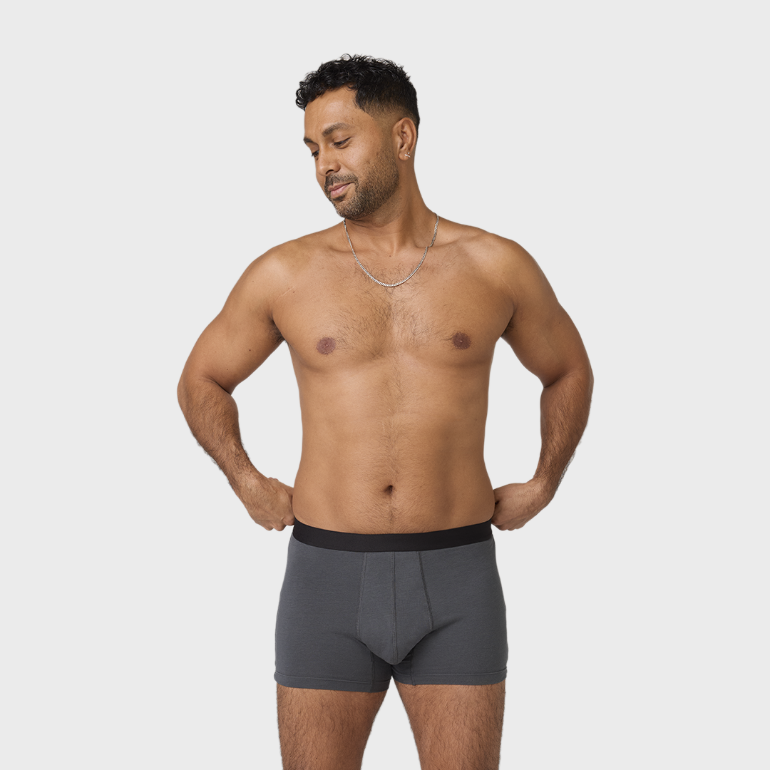 Man wearing NEW TENA Men Protective Boxers, adjusting charcoal-gray boxer briefs with black waistband, showcasing stylish design and comfort against a plain background.