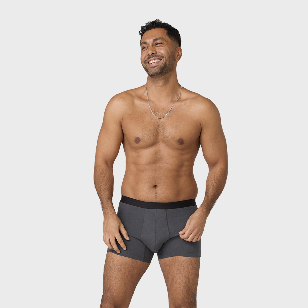 Man wearing NEW TENA Men Protective Boxers and a silver chain necklace smiles, showcasing the boxers' soft, organic cotton material and stylish design.