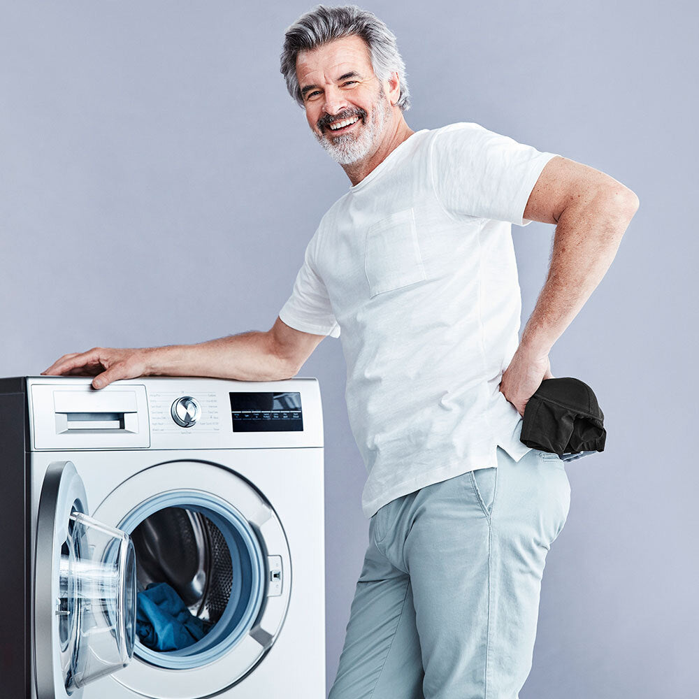 Man with a pair of black men's washable incontinence boxers in his hand, smiling and leaning on a front-loading washing machine on a grey background.