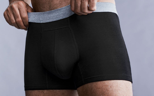 Male hands adjusting men's black washable incontinence boxers with a grey waistband in front of a grey background.